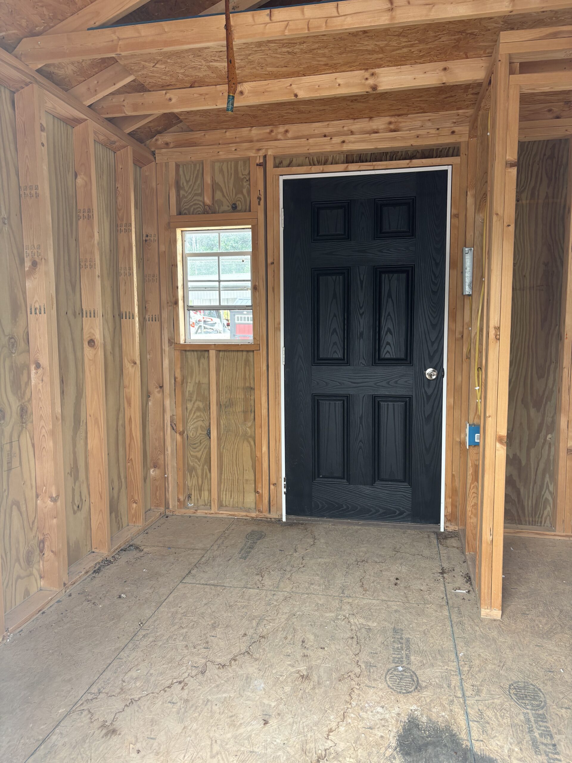 Unfinished interior with exposed wooden framing, small window to the left, and a black paneled front door.