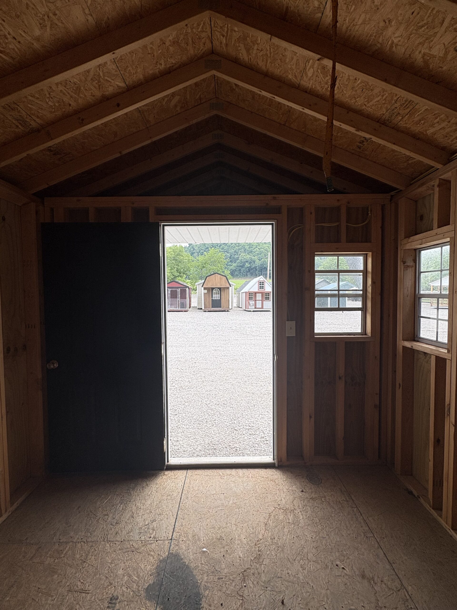 Open doorway to a gravel yard with small colorful sheds visible outside; unfinished wooden interior framing and exposed OSB ceiling surround the opening.