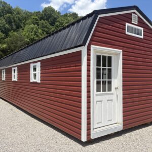 Red metal-siding shed with white-framed door on a gravel lot and green trees in the background.