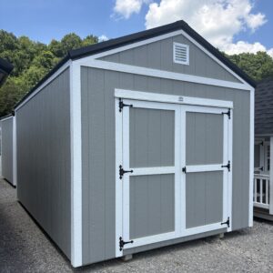Gray storage shed with white trim and double doors on a gravel surface, under a blue sky with clouds, and a vent on the gable.
