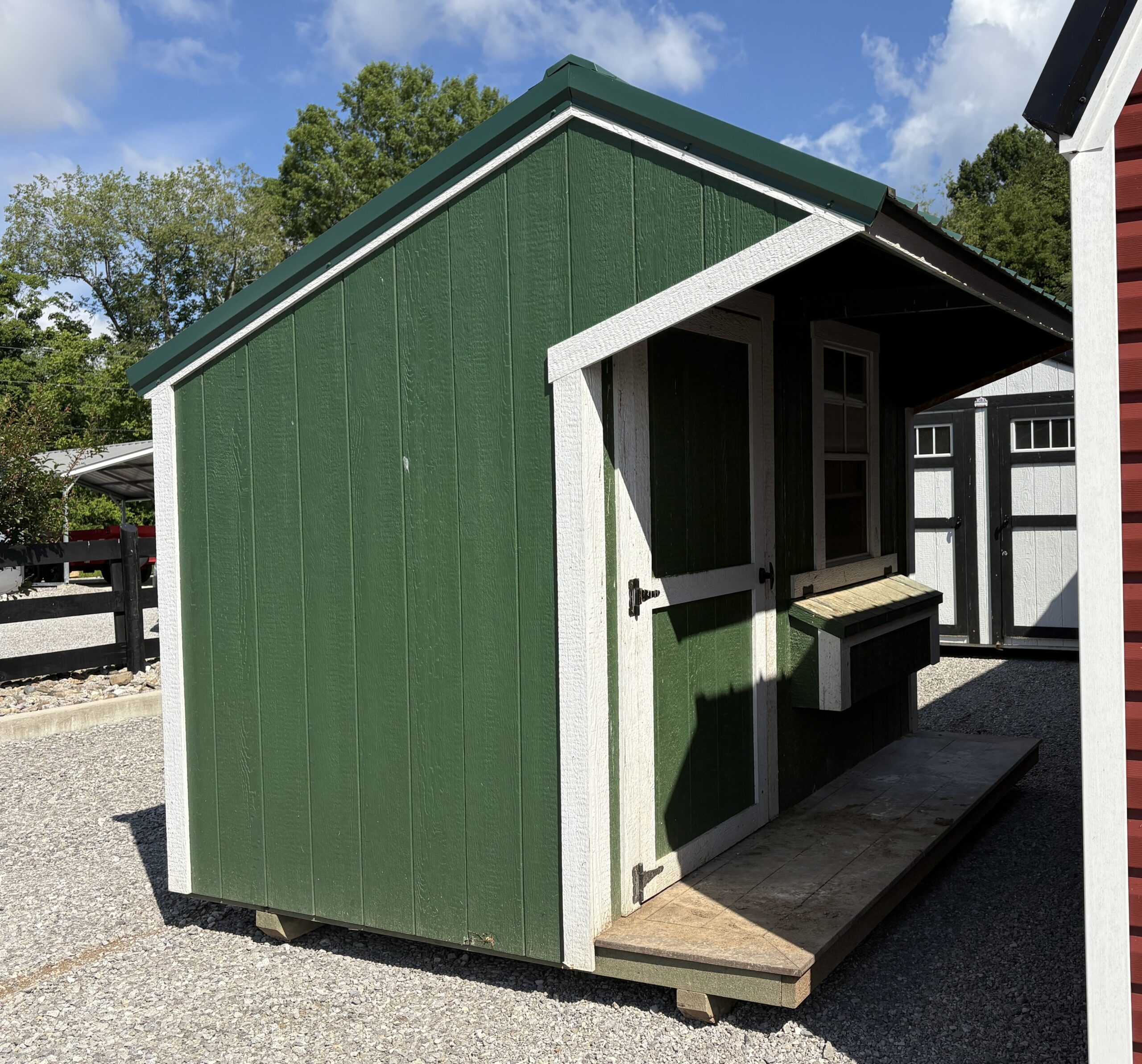 Green wooden shed with white trim and an open service window, set on gravel under a blue sky with trees in the background with a sloped roof.