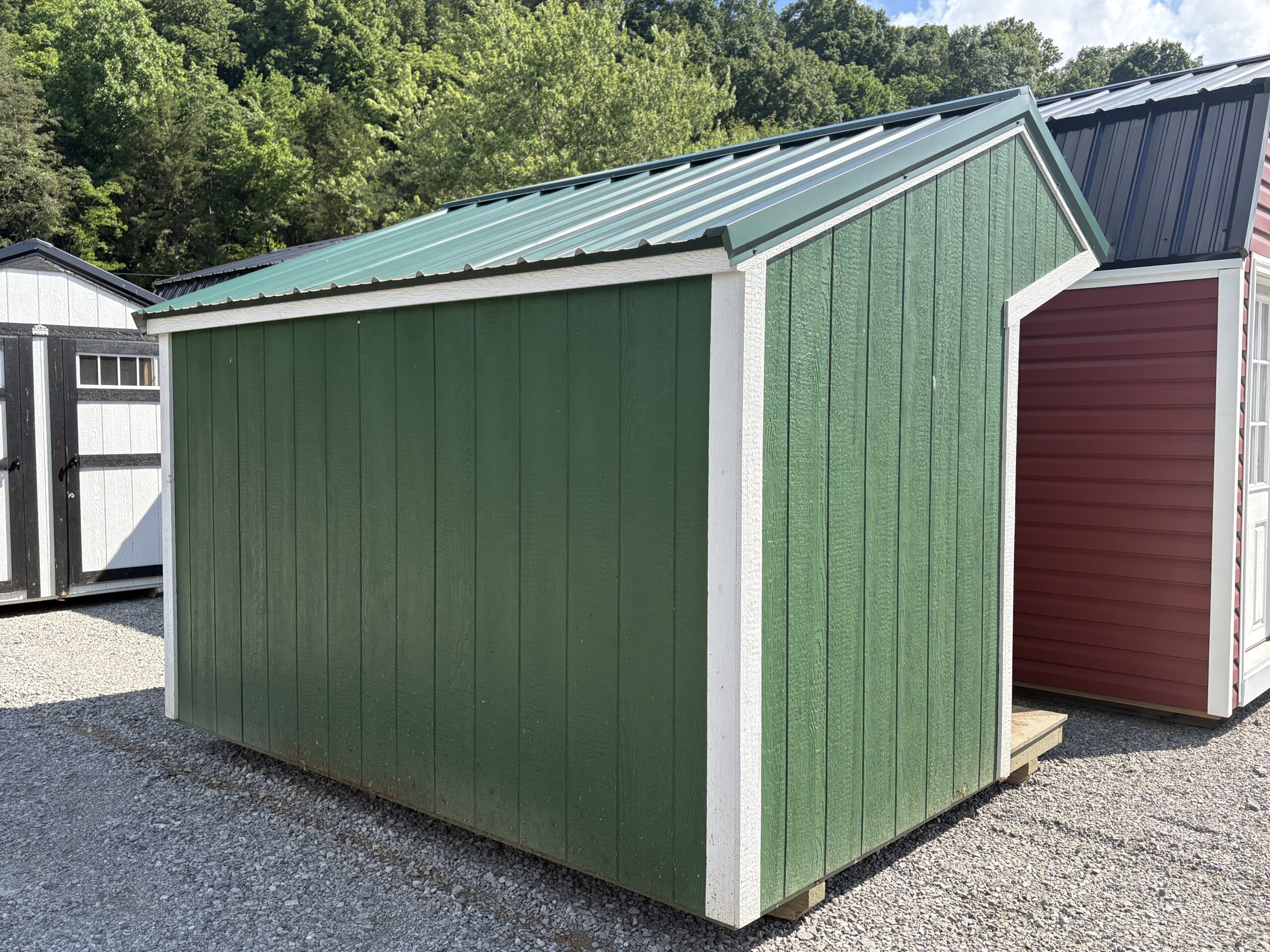 Green wooden storage shed with white trim and a green metal pitched roof in a gravel yard, trees in the background