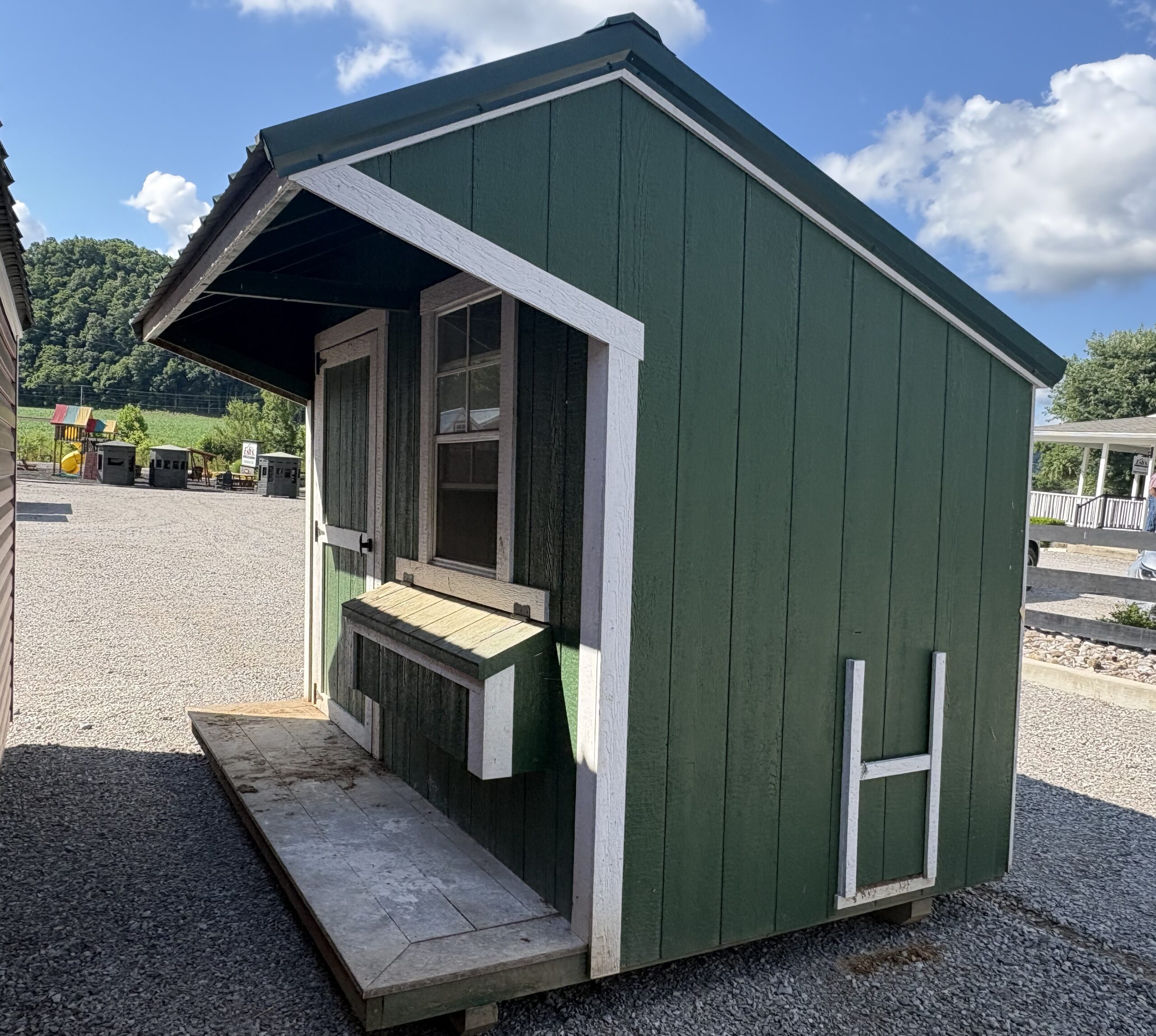 Small green wooden shed with white trim and a counter window on a gravel lot, with a playground in the background.
