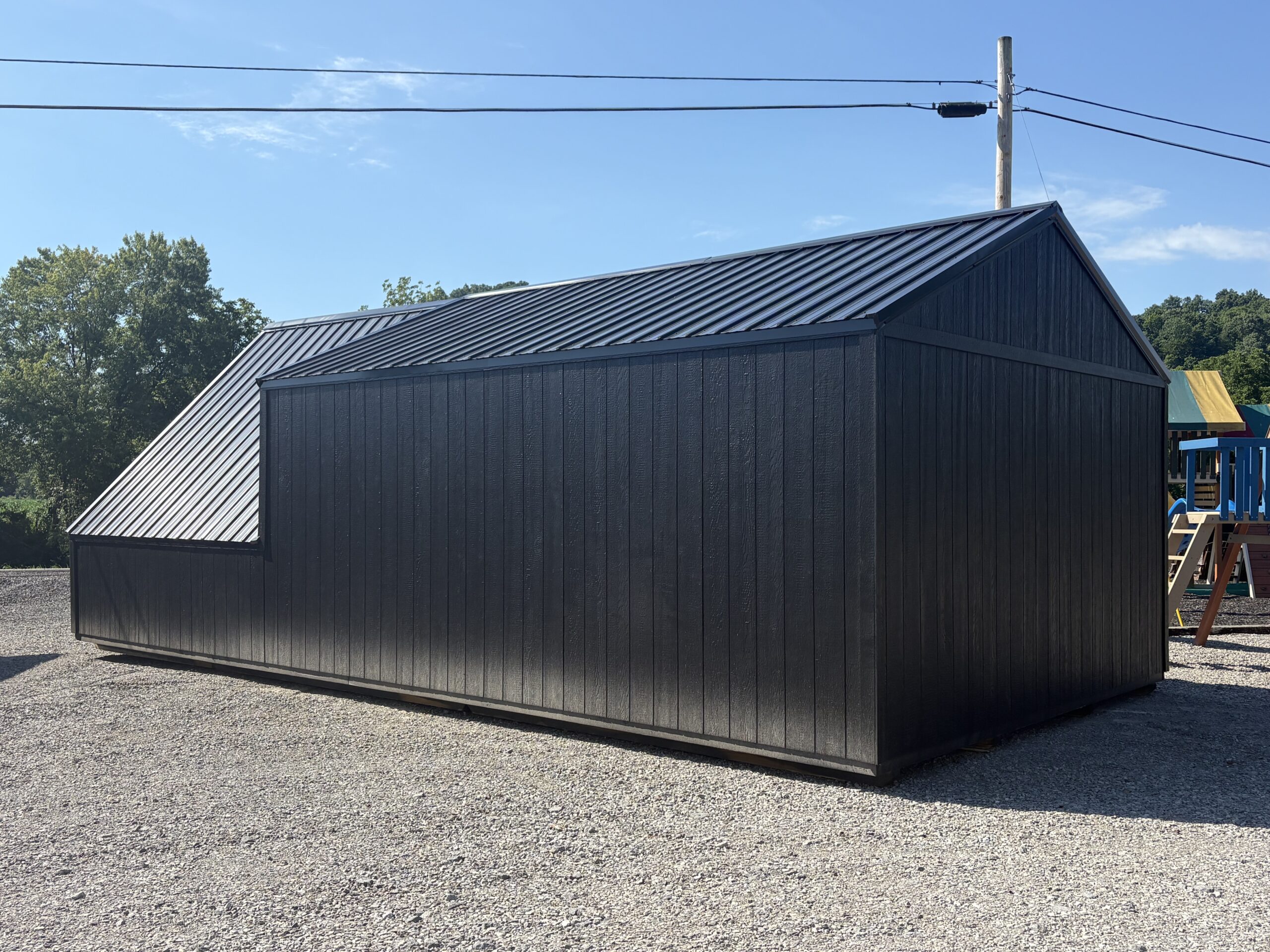 Gray metal storage shed with a steep, sloped roof in a gravel lot, under a blue sky with power lines above.