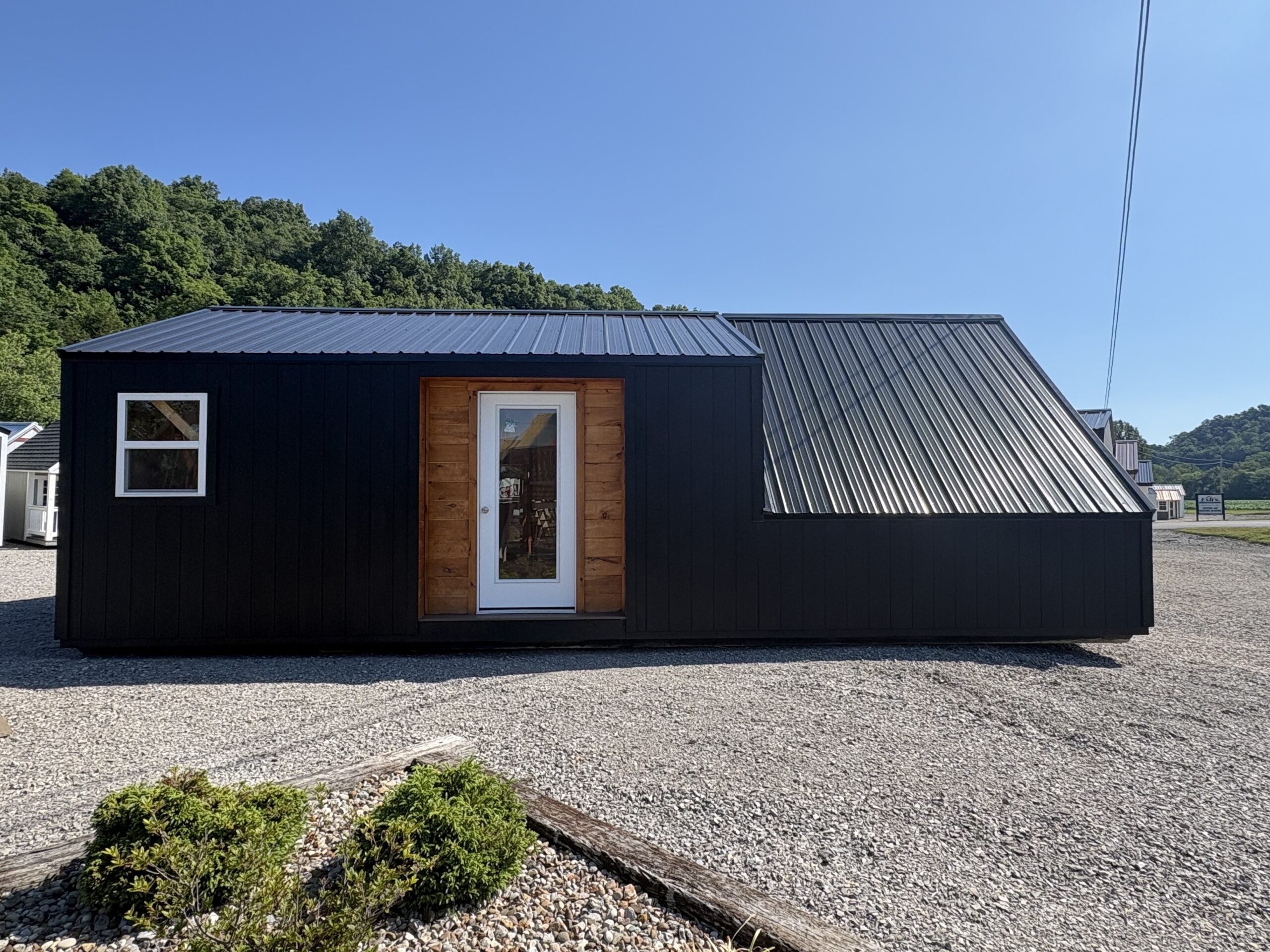 Front view of a black modular building with a wooden entry door and white-framed window, metal roof, on a gravel lot with a hillside in the background.