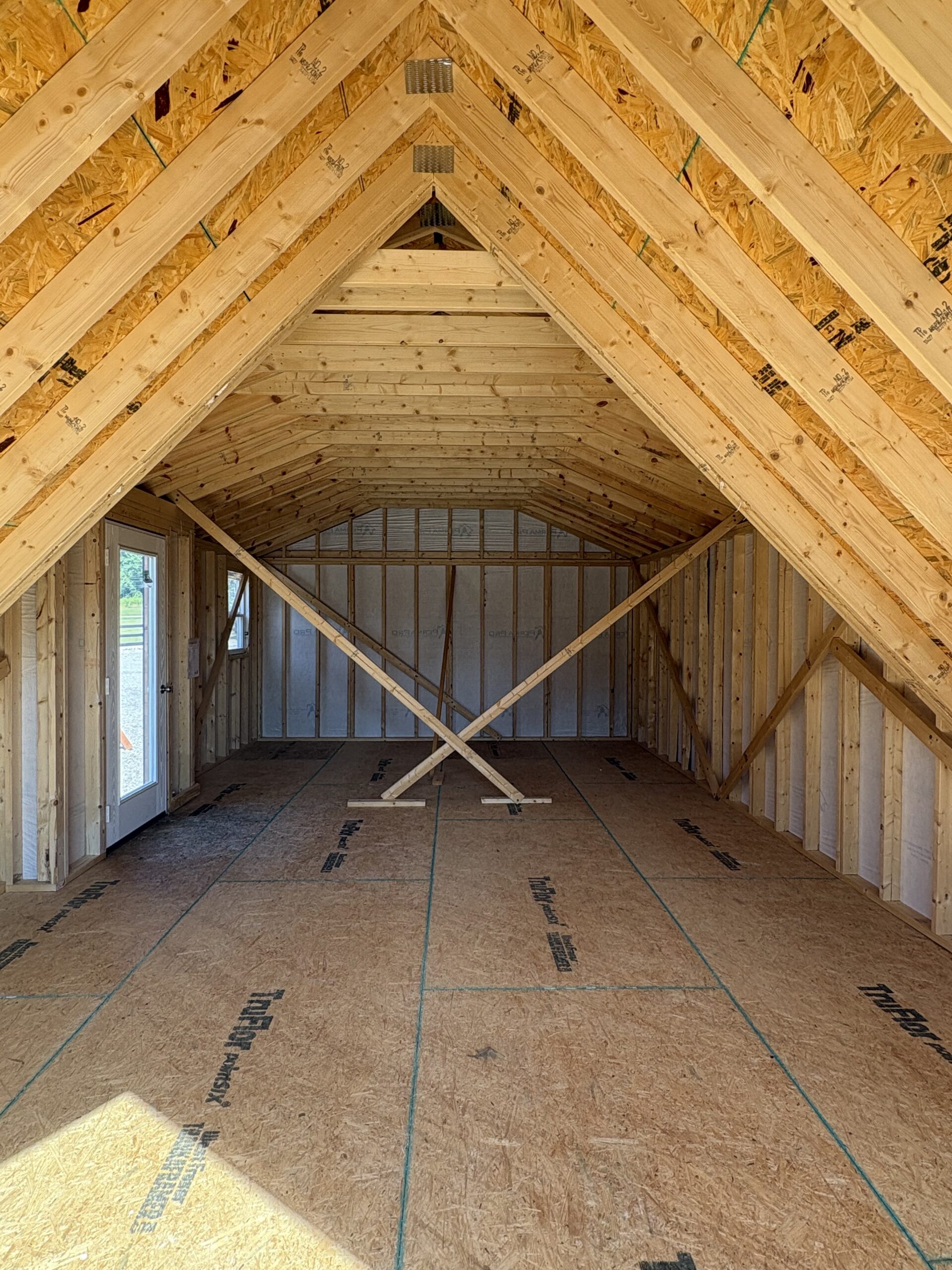 Unfinished attic with exposed wooden roof trusses forming a central peak and cross braces; plywood walls and floor panels visible, with a door on the left.