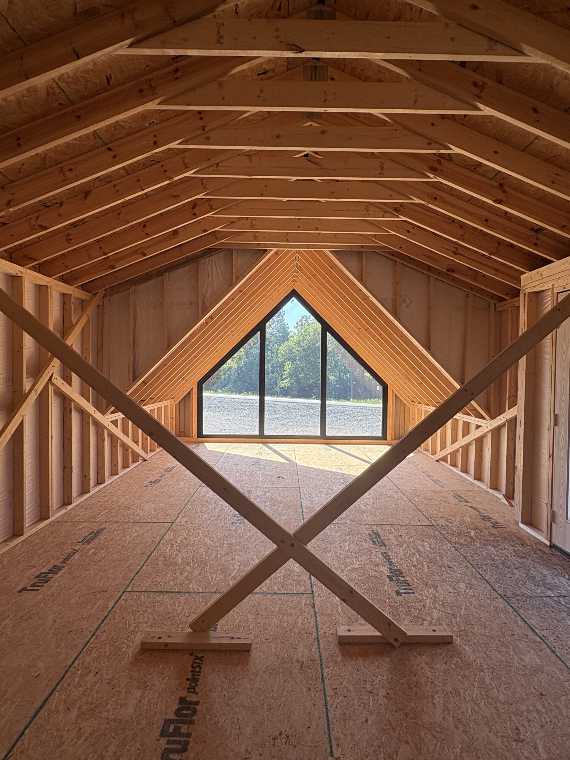 Attic under construction with exposed wooden roof trusses and a large triangular window at the far end, letting in daylight over trees.
