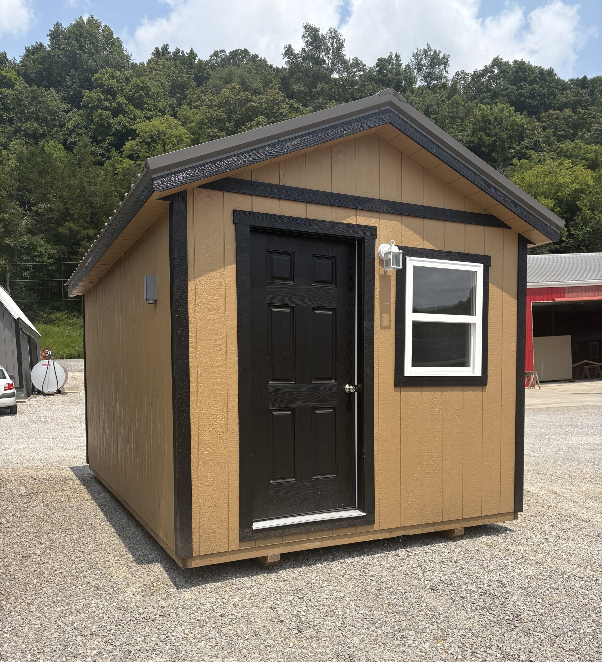 Small tan shed with black door and trim, white-framed window, outdoor light, on a gravel lot with trees in the background