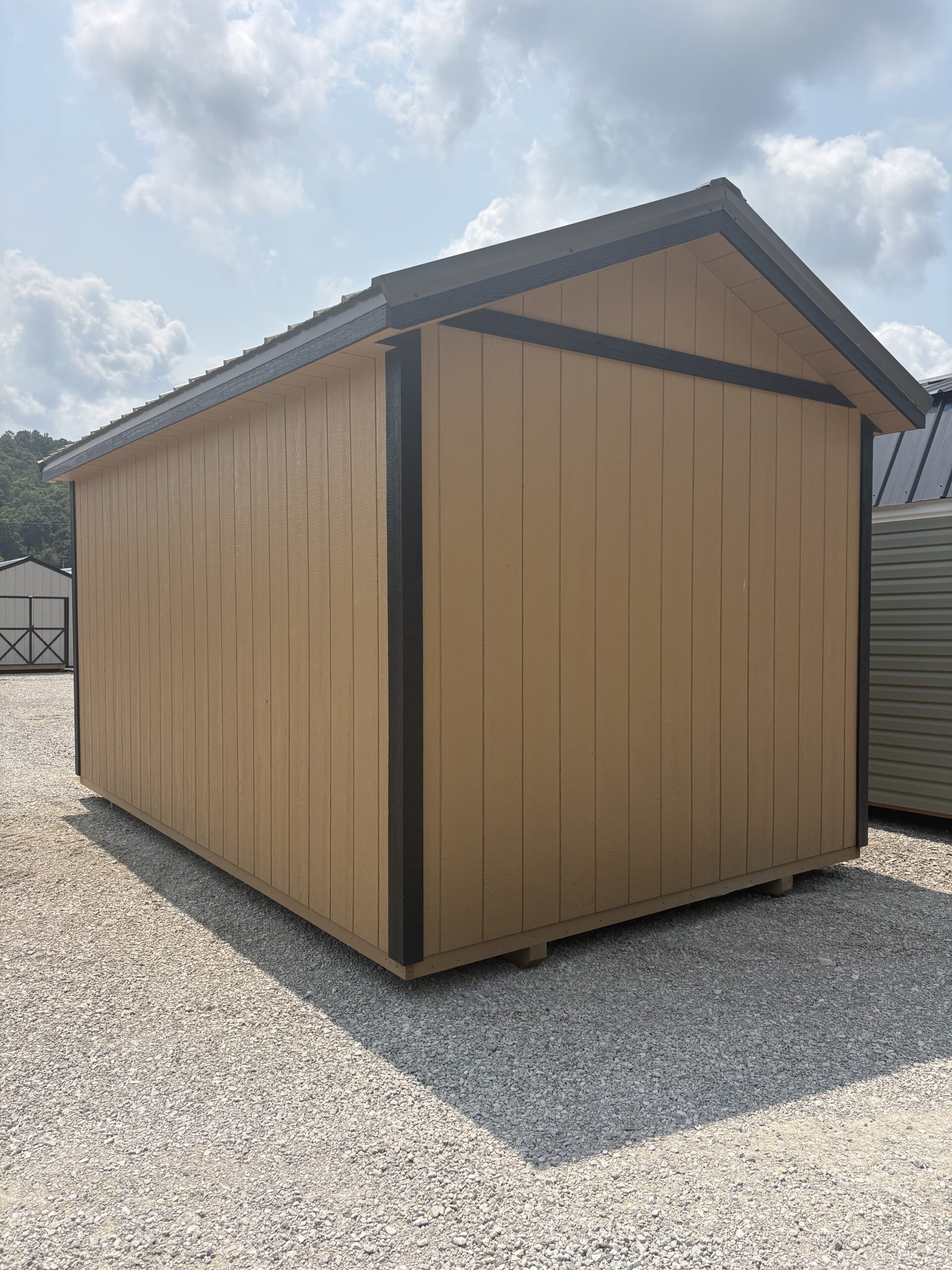 Tan wooden shed on a gravel lot with dark trim and a sloped roof against a cloudy sky