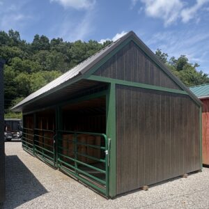 Wooden barn-like structure with dark siding and green trim, open side, metal roof, on gravel lot with forested hill behind it.