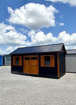Dark blue shed with warm wooden doors and window frames under a bright blue sky with fluffy clouds in a gravel lot, outdoor display setting.