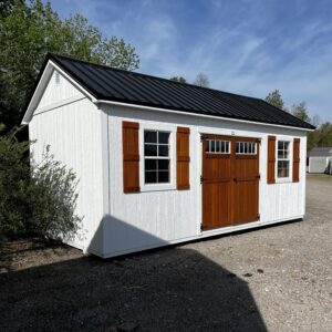 White wooden shed with brown shutters and a double barn-style door on a gravel lot under a blue sky.