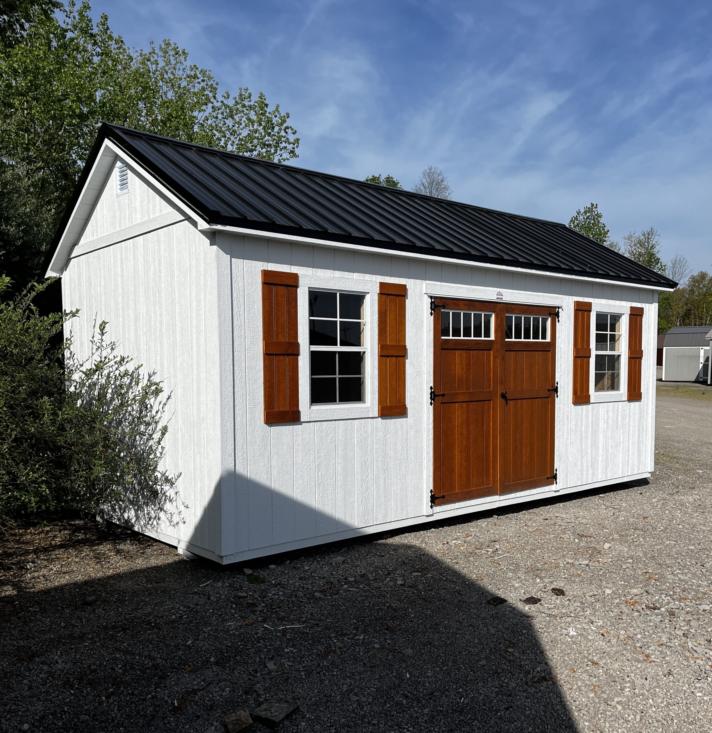 White wooden shed with brown shutters and a double barn-style door on a gravel lot under a blue sky.