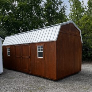 Brown wooden shed with a silver corrugated metal roof, set on a gravel lot, with green trees in the background