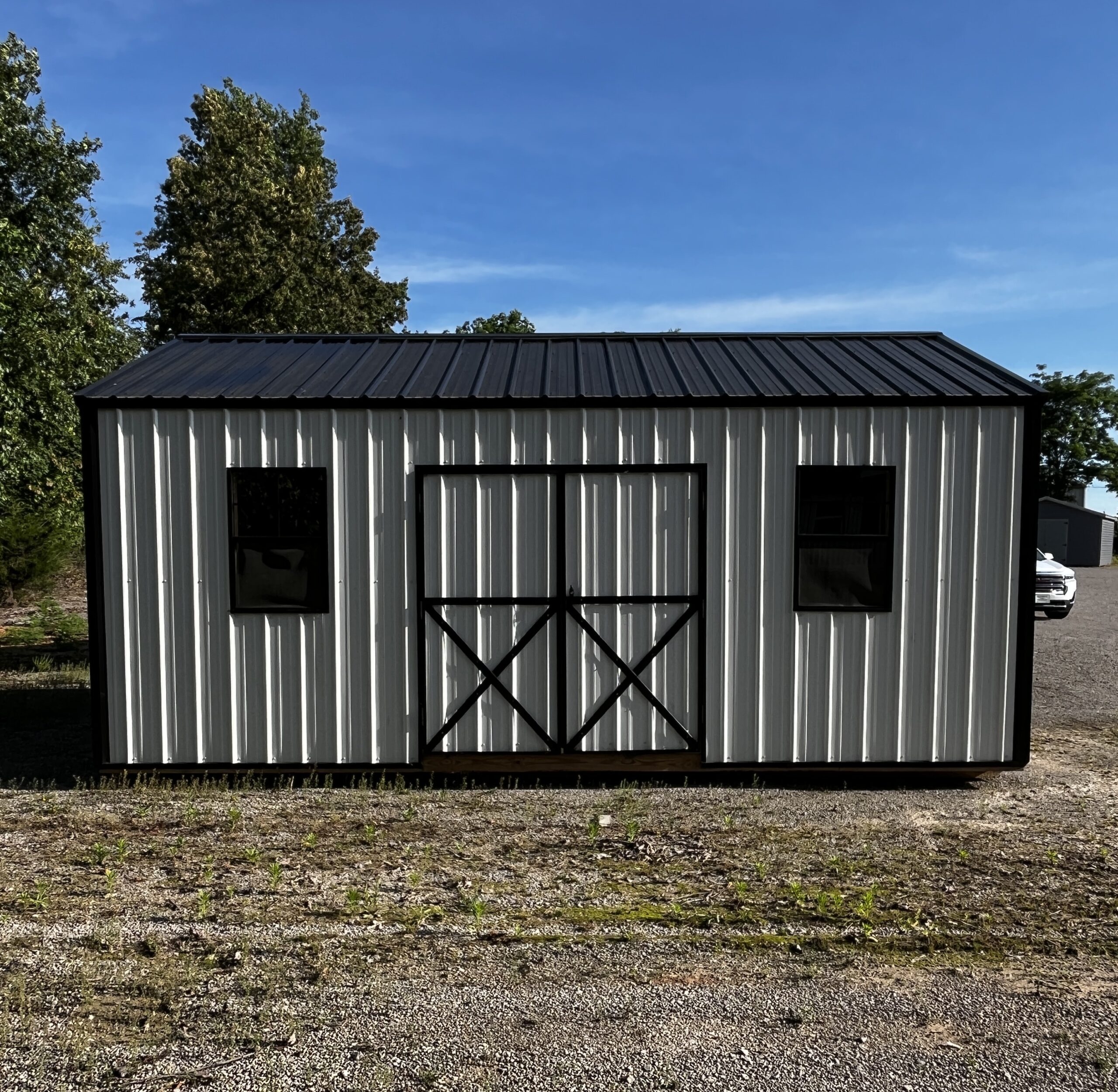 Metal shed with vertical corrugated panels and black-framed doors, set on a gravel lot beneath a blue sky with trees nearby.