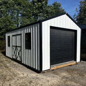 White corrugated metal shed with black trim and a large dark garage door, placed on wooden pallets in a gravel lot with trees and blue sky behind.