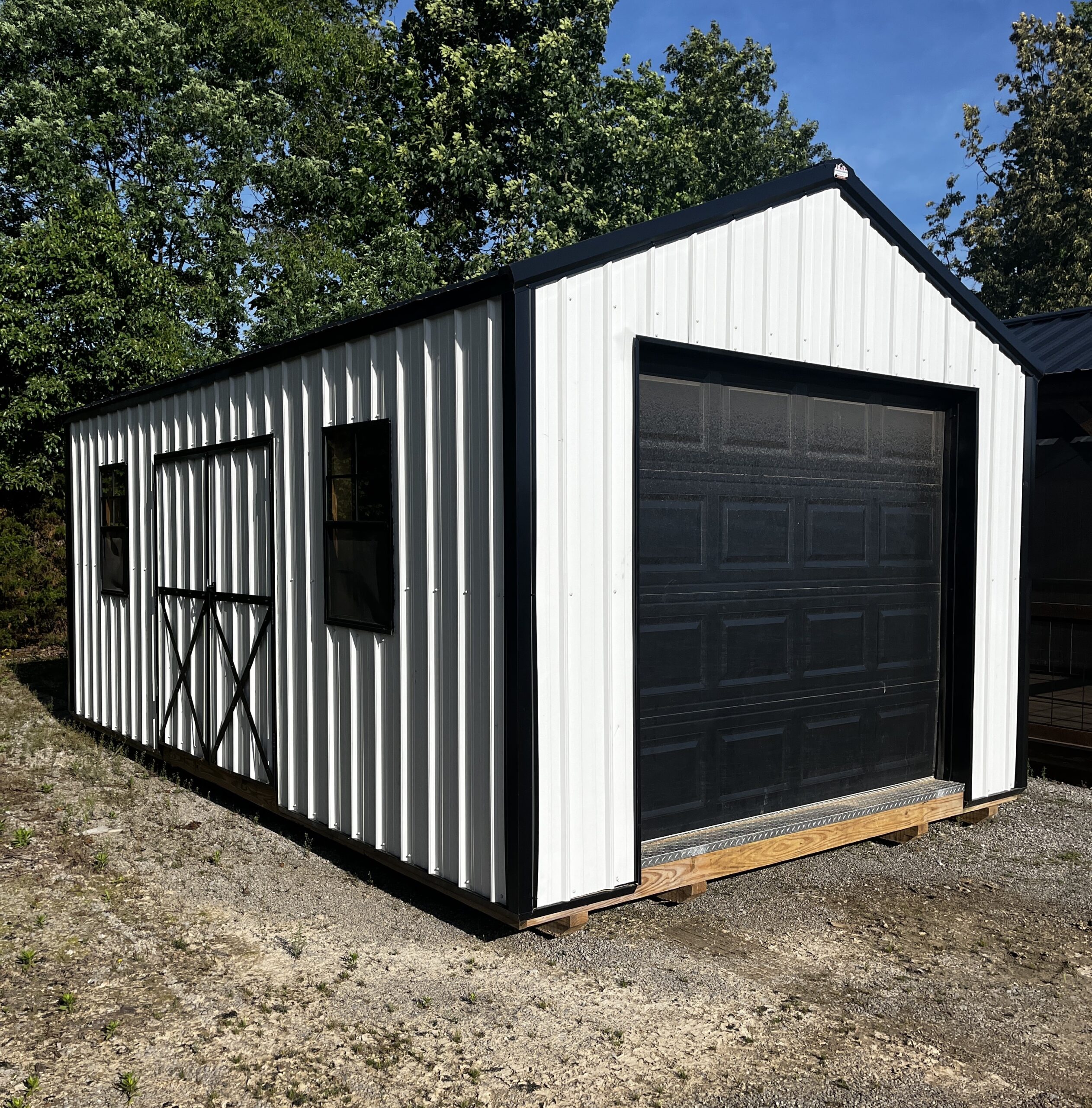White corrugated metal shed with black trim and a large dark garage door, placed on wooden pallets in a gravel lot with trees and blue sky behind.