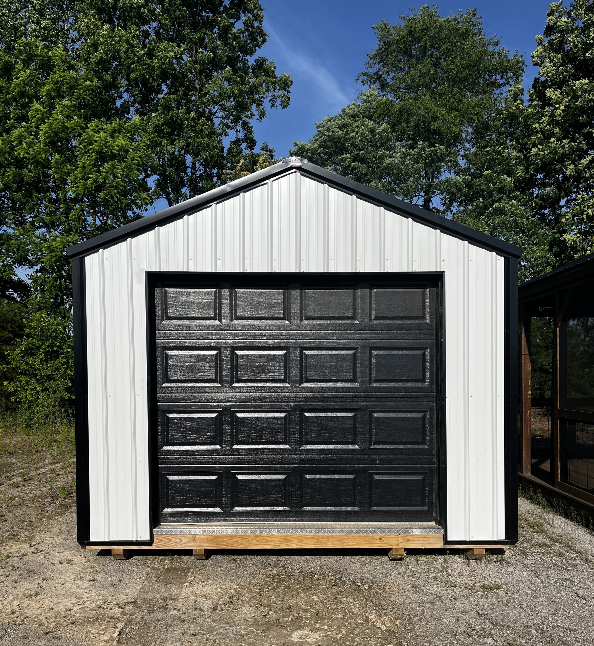 Black raised-panel garage door set in a white metal-framed building, resting on a wooden pallet outdoors with trees and blue sky in the background
