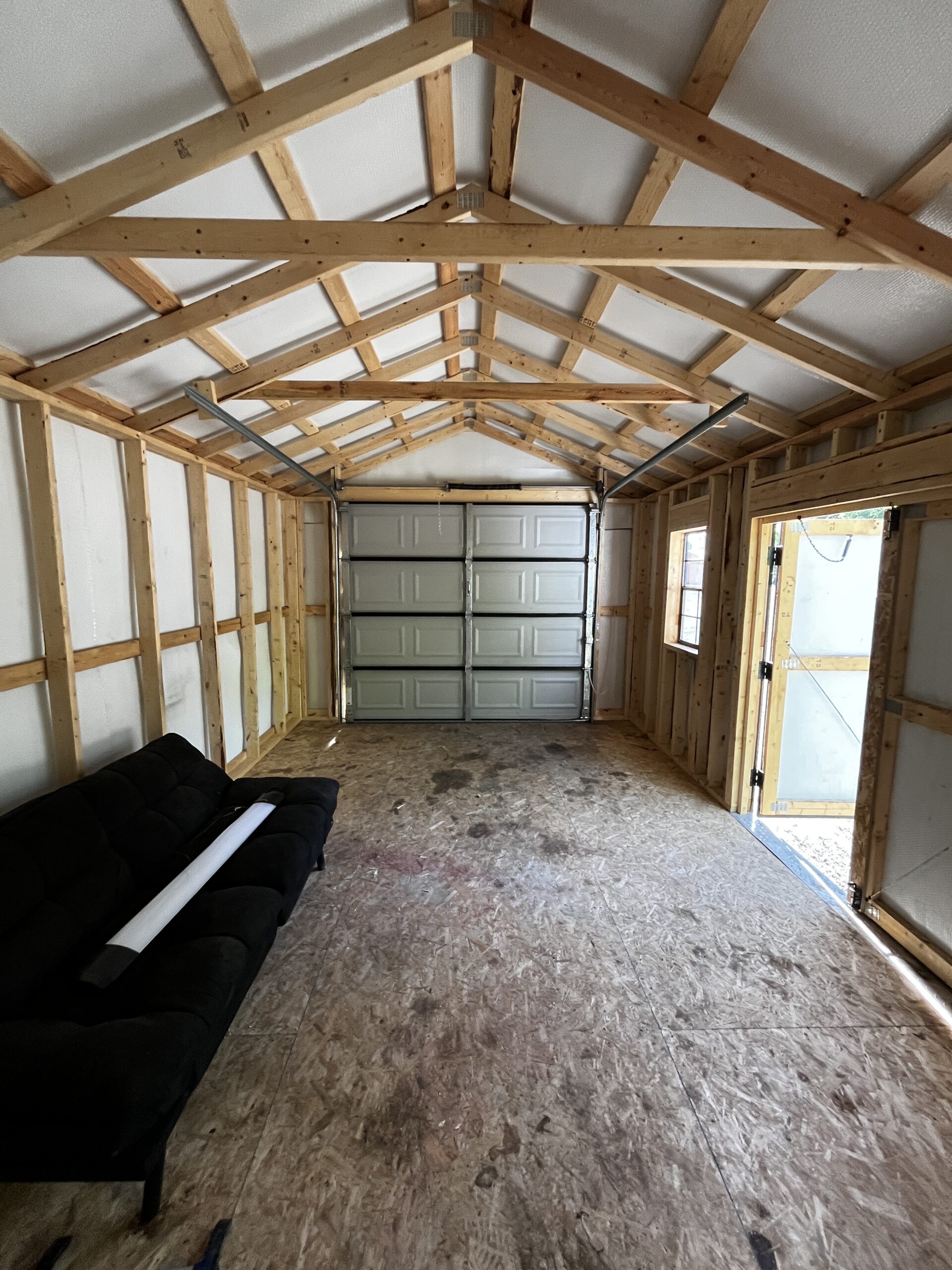 Unfinished garage with exposed wooden framing and trusses, a gray/black sofa along the left wall, and a metal sectional garage door at the far end.
