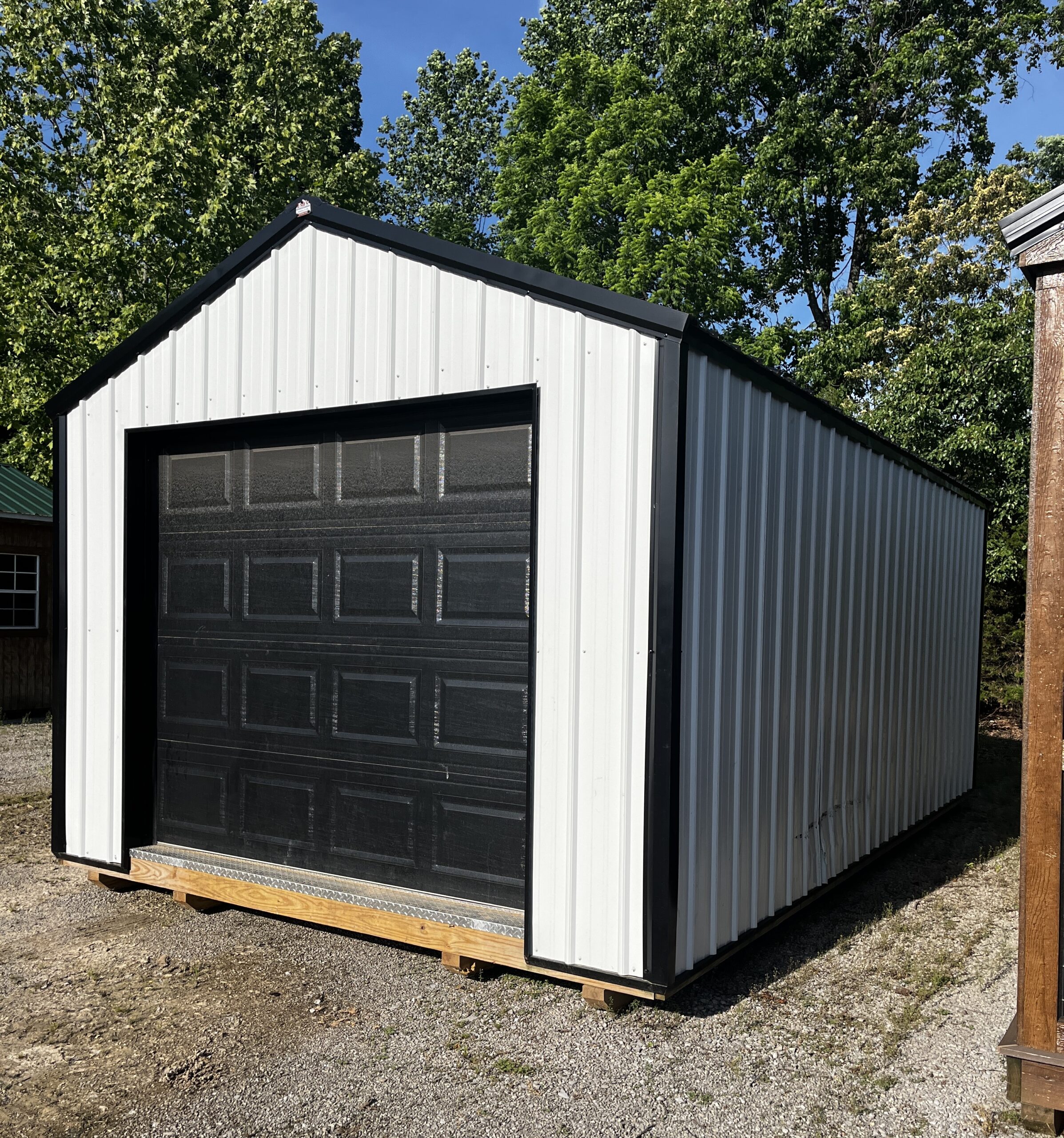 Metal storage shed with a large black garage door on a wooden pallet in a gravel yard, with green trees and blue sky behind.