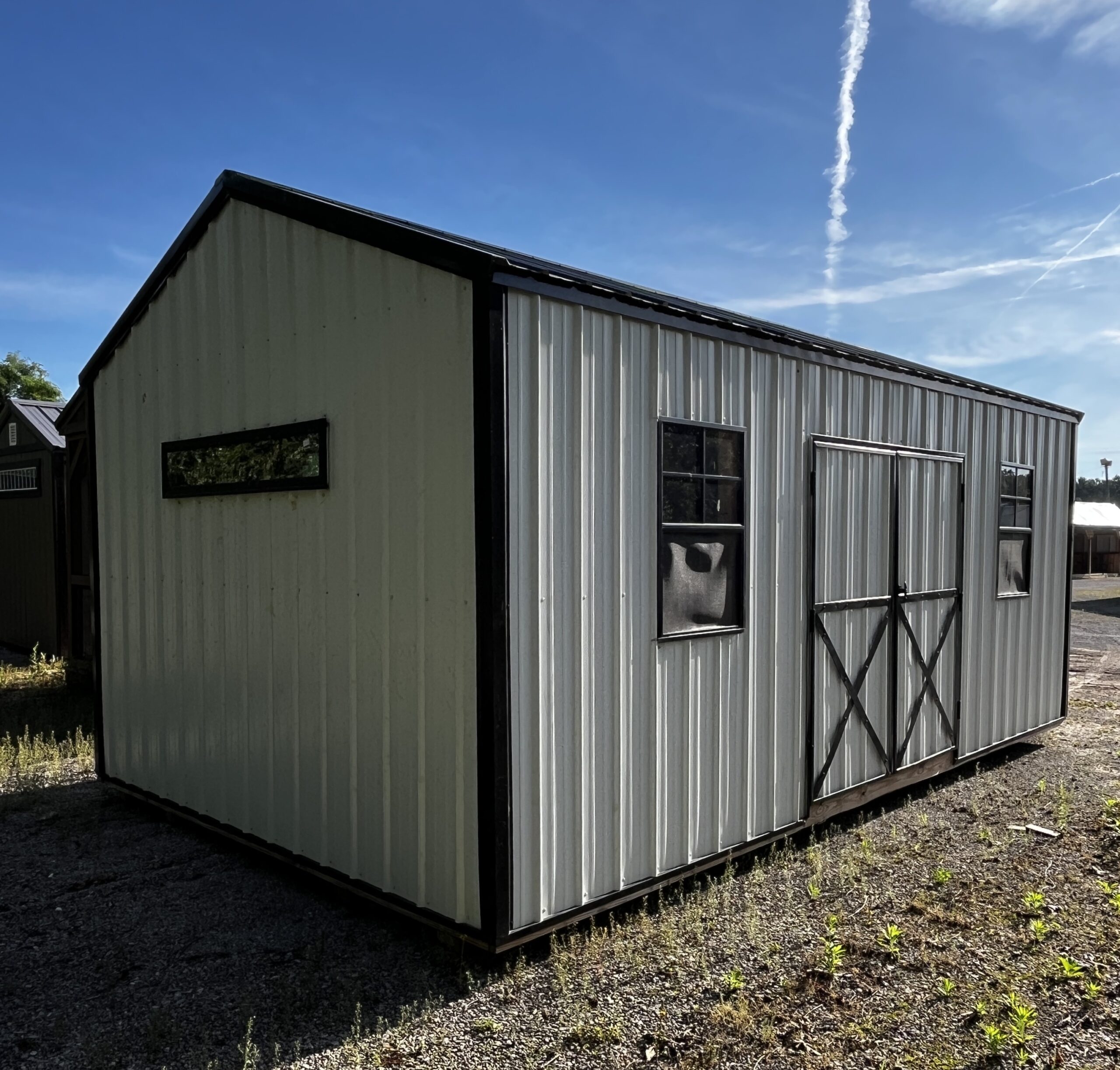 Corner view of a light-gray corrugated metal shed with black trim on a gravel lot under a clear blue sky and a white contrail high in the air.