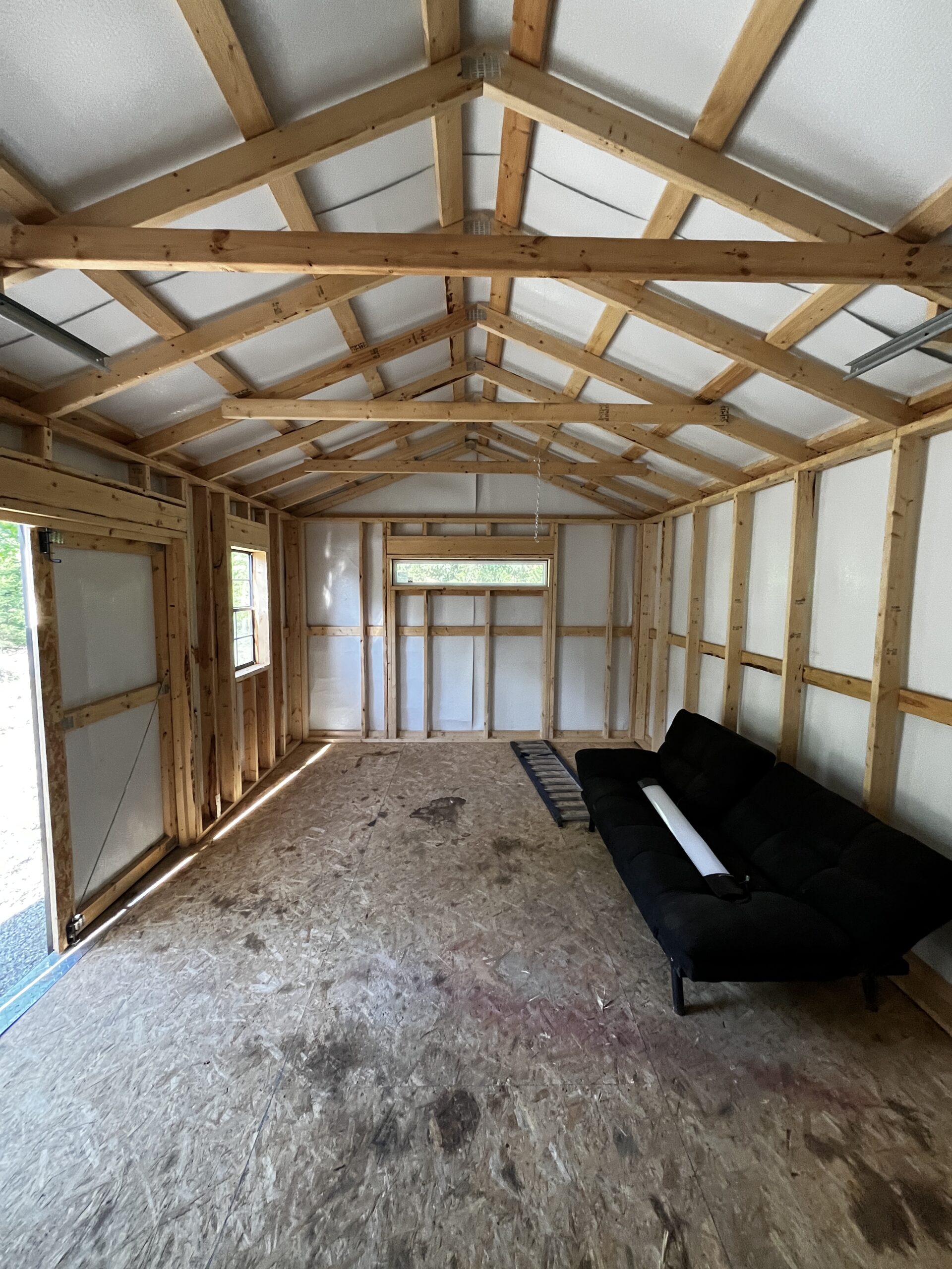 Interior of an unfinished framed building with exposed wooden studs and roof trusses; a ladder and a black sofa on the floor.