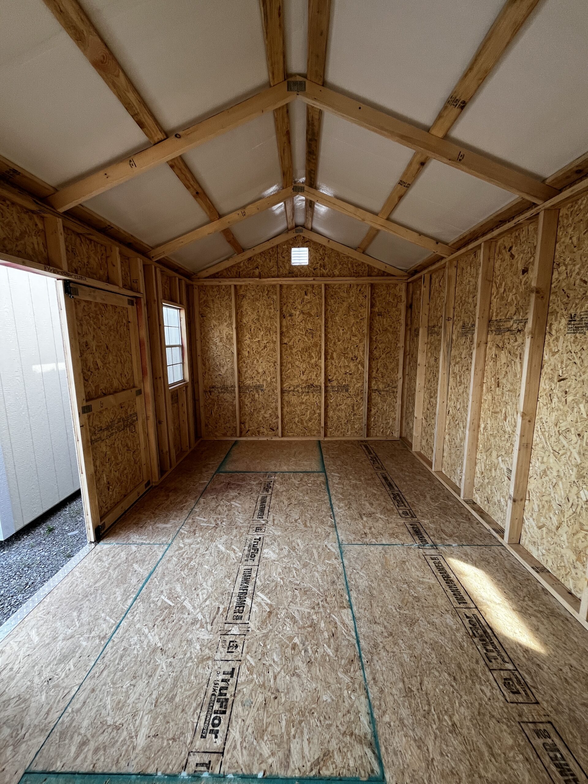 Interior of an unfinished wooden shed with exposed studs, roof trusses, and OSB walls; a small vent at the far end.