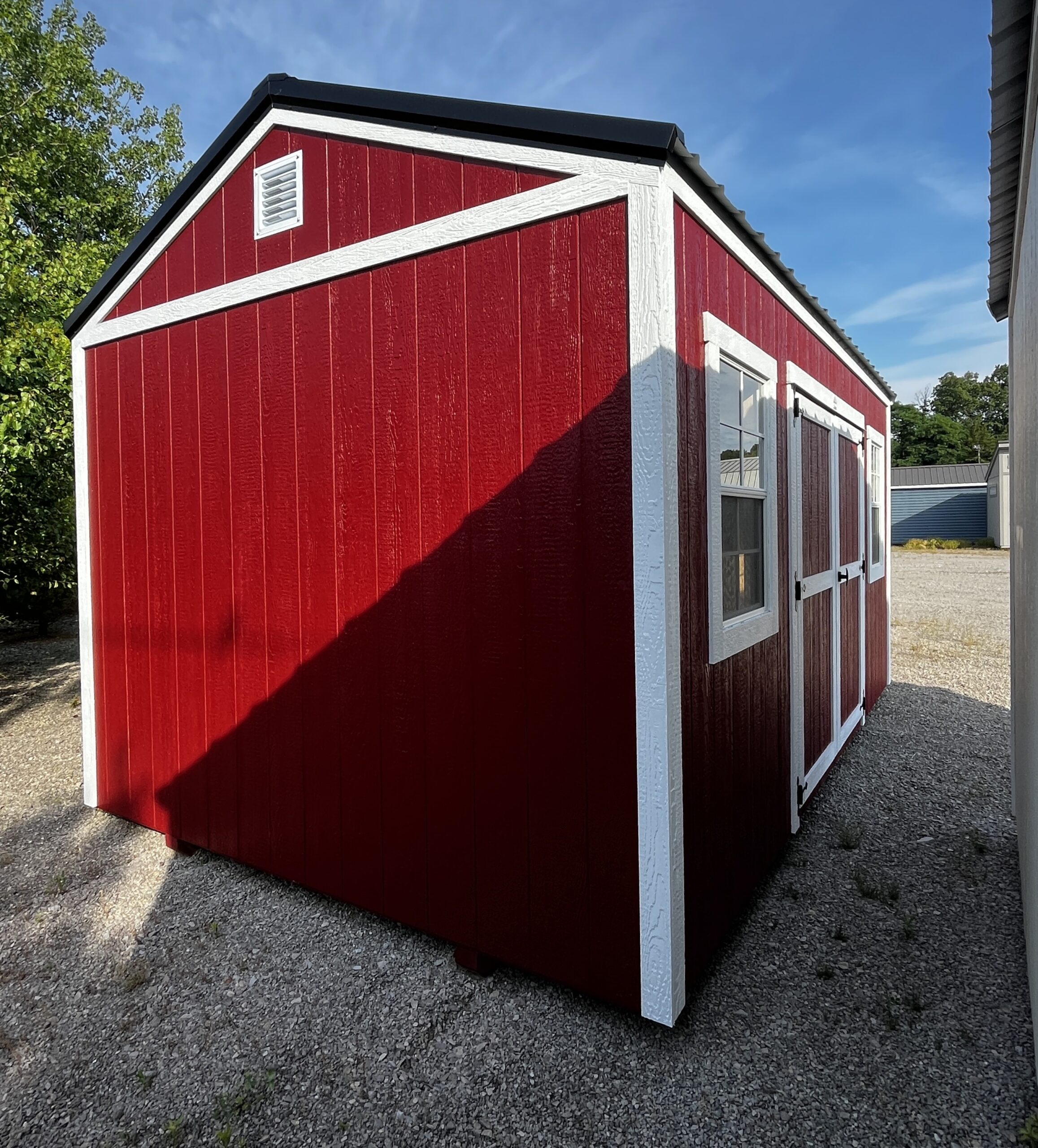 Red wooden shed with white trim and diagonal white trim accent, blue sky above, gravel ground surrounding the structure, small vent near the peak.