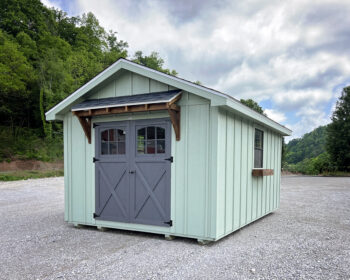 Mint-green shed with gray double doors on a gravel lot, trees in the background and a cloudy sky above.