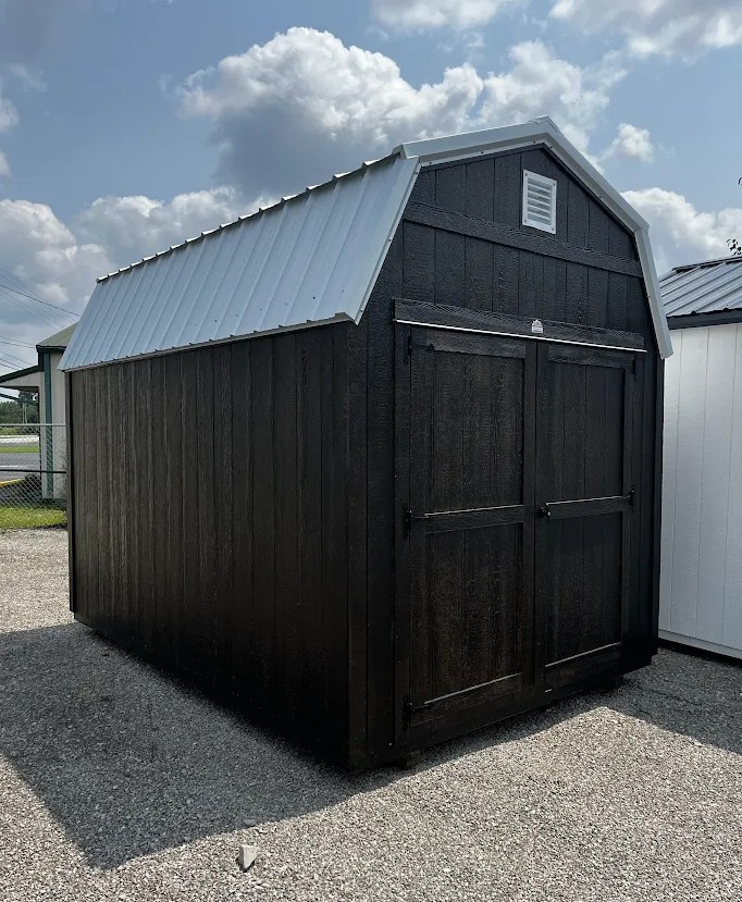 Black wooden storage shed with a light gray metal pitched roof on a gravel lot, featuring double barn doors and a small vent near the peak.
