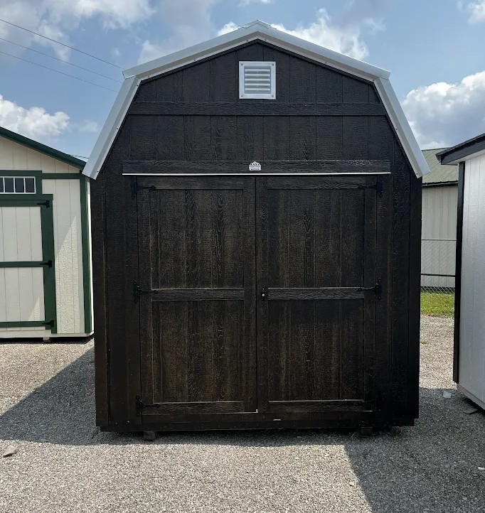 Black wooden shed with double doors and a small vent near the peaked roof, set on a gravel lot beside other storage sheds.