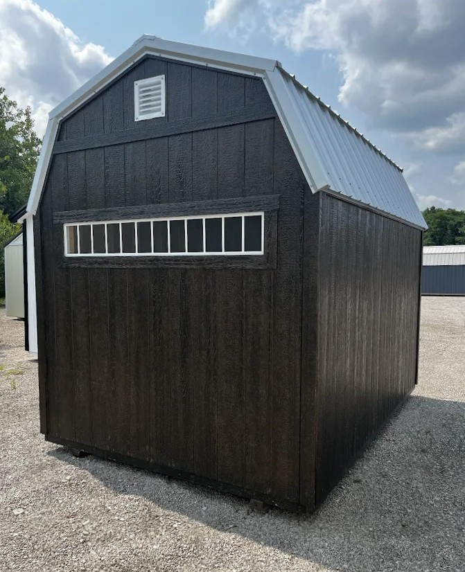 Dark wood storage shed with a pale gray metal roof, vent at the gable, and a long horizontal window with bars, on a gravel surface.