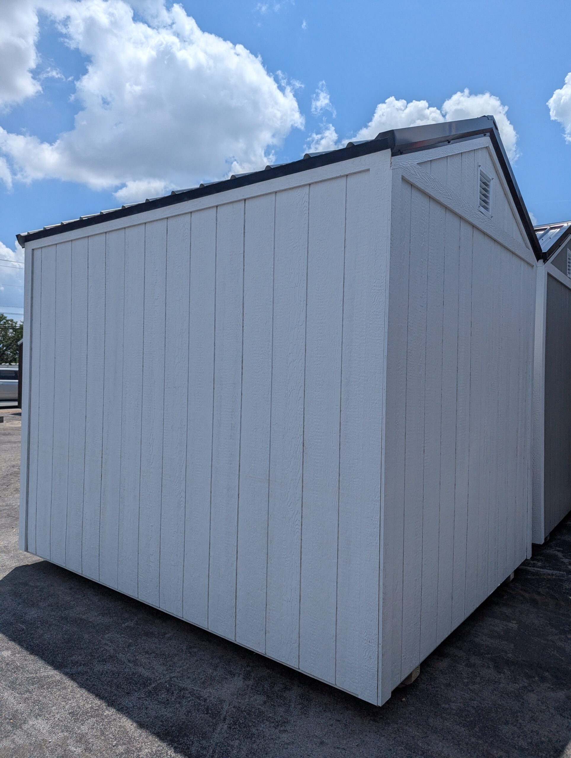 White wooden storage shed with vertical siding on a concrete surface, under a blue sky with clouds.