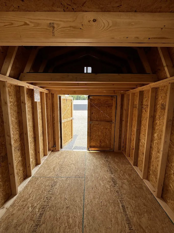 Interior of a wooden-framed shed under construction, with plywood walls, exposed studs, and an open door to the outside light pale beyond the doorway.
