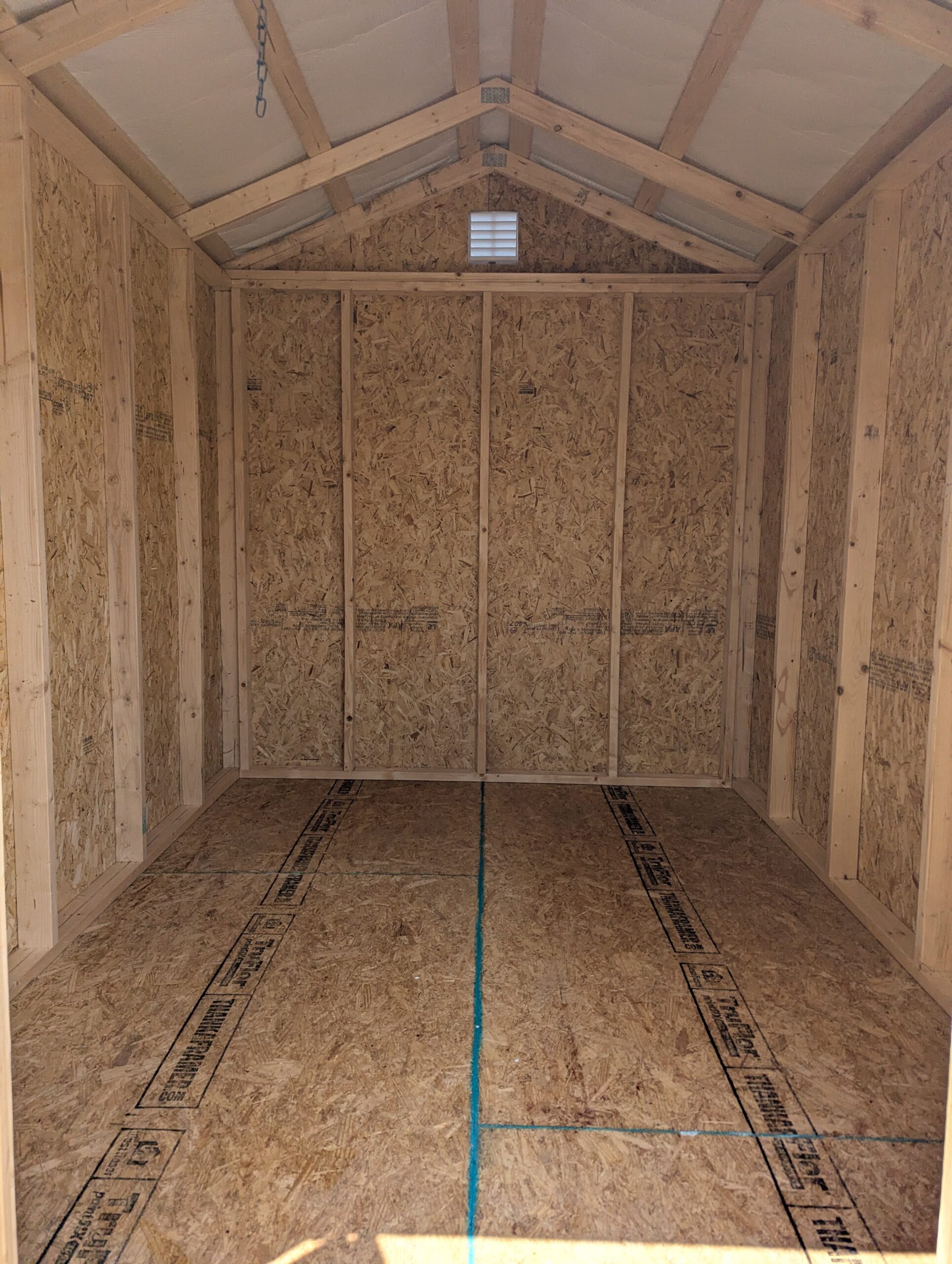 Unfinished wooden shed or attic with exposed stud framing and OSB walls, triangular roof trusses, a small vent, and a blue line drawn along the floor for layout.