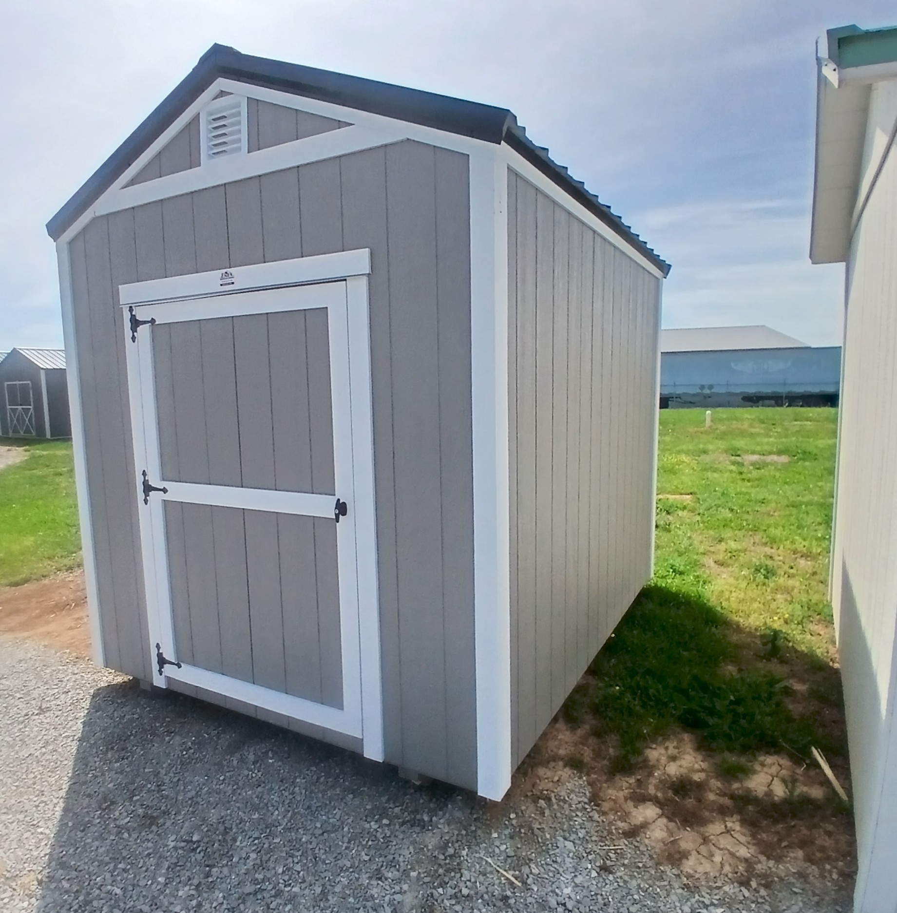 ng - Esh's Utility Buildings Gray wooden storage shed with white trim and a peaked roof, on a gravel surface beside a grassy area.