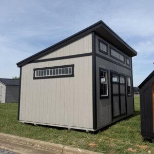 Two-tone shed with a tall slanted roof, beige walls and black trim, windows along the front and side on a grassy lot.