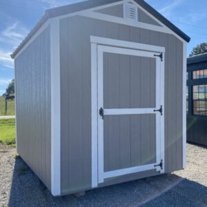 Small gray storage shed with white trim and a framed single door, siting on gravel under a blue sky with a vented gable.