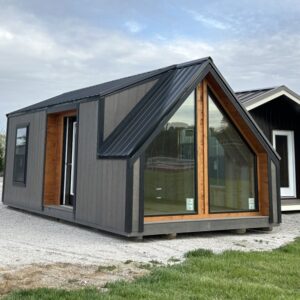 Dark gray prefab tiny house with a steep, metal roof and a large glass front wall framed in wood.