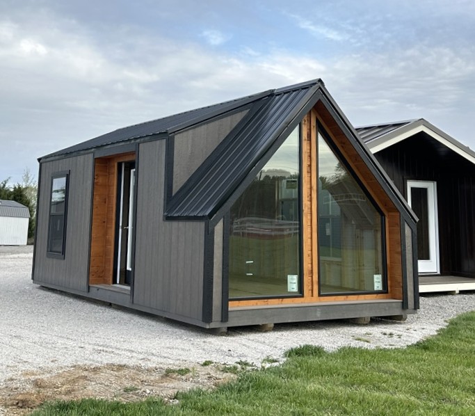 Dark gray prefab tiny house with a steep, metal roof and a large glass front wall framed in wood.