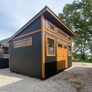 A small black wooden shed with orange trim and multiple windows on a gravel lot, featuring a double door entry.