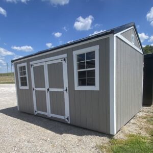 Gray garden shed with white trim, double barn doors, and a window on the side, set on gravel under a blue sky.
