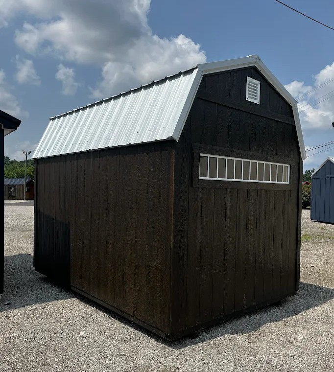 Small dark brown shed with a light metal roof on a gravel lot, featuring a long row of narrow rectangular windows on the side and a small vent near the peak.